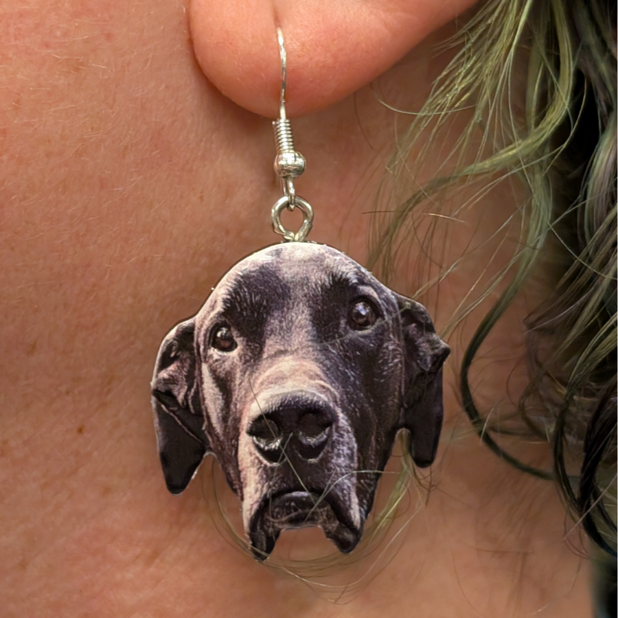 A close-up of a person's ear wearing a dangling earring shaped like a black dog's face. The earring has a silver fishhook back and a detailed dog head charm.