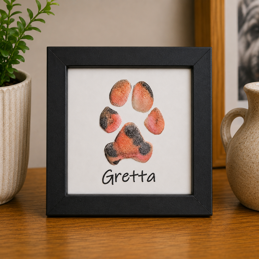Framed print of a dog paw print with 'Gretta' on a table next to a plant and vase.